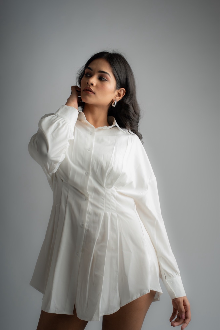 Hand raised behind her neck, Shreya tilts her head in a white shirt dress against a soft grey wall
