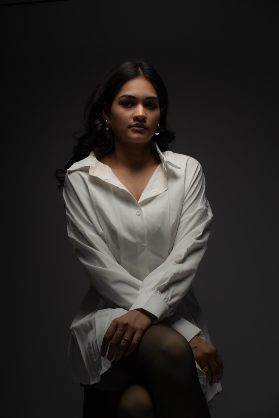 Low-key frame of Shreya perched on a stool in a white shirt and dark tights, near-black backdrop