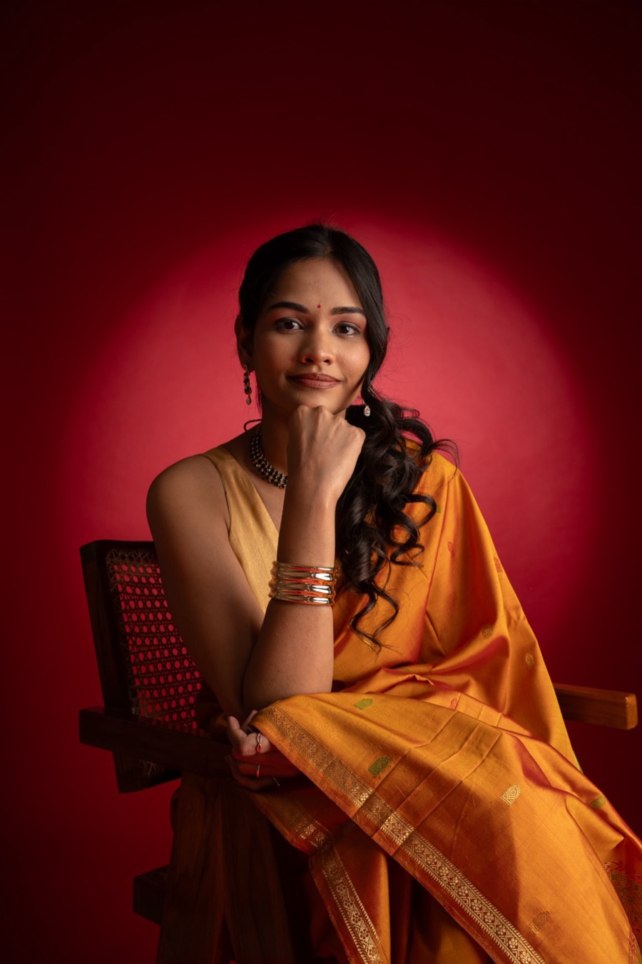 Shreya in a mustard silk saree and gold bangles, chin on her hand, seated against a deep red backdrop