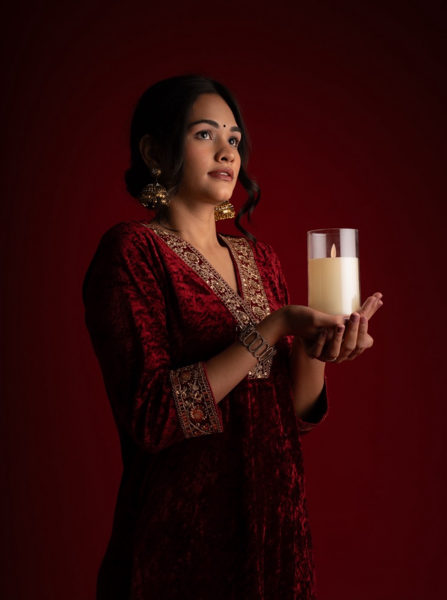 Holding a lit candle in glass, Shreya wears a deep red embroidered kurta against a matching red wall