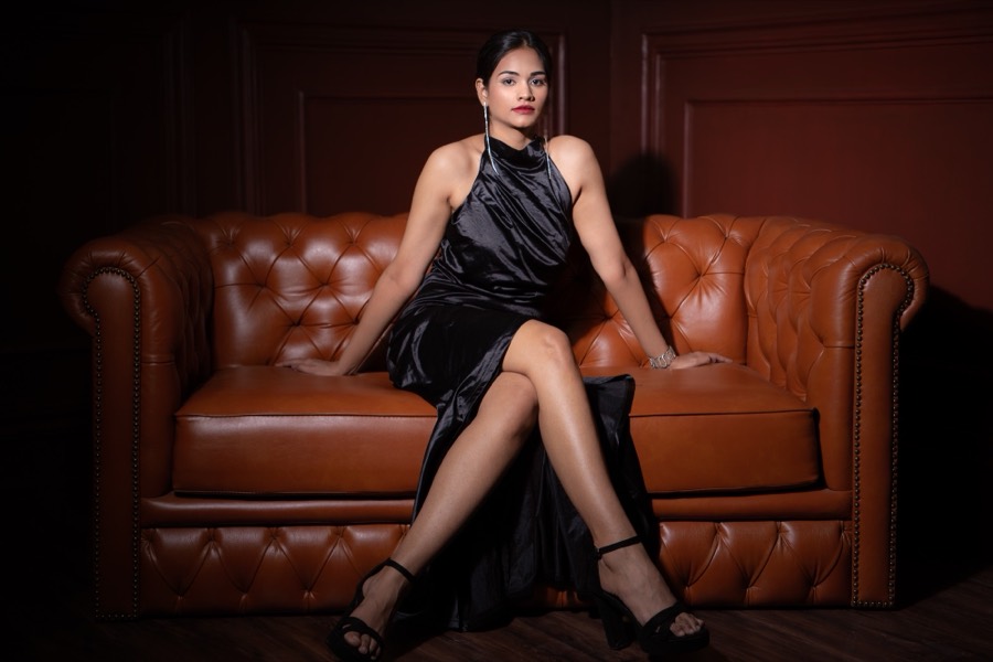On a tan leather Chesterfield in a black satin gown and heels, Shreya crosses her legs in a dark room