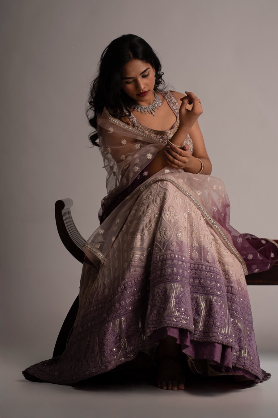 Looking down at her hands, Shreya wears a lilac ombre lehenga and sheer dupatta on a cane chair